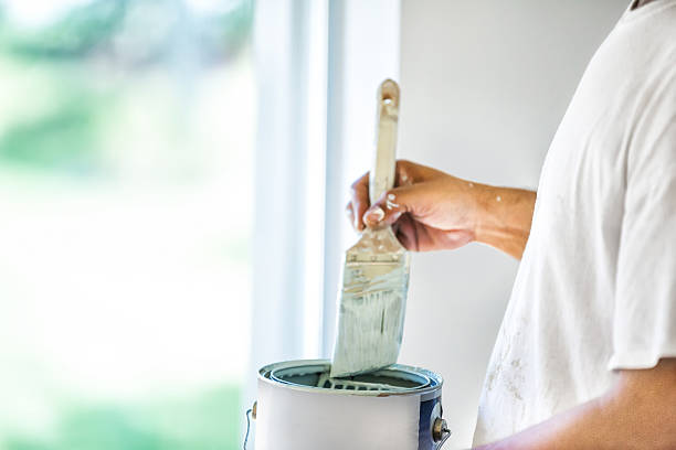 A house painter dips his brush into a can of paint.  rr